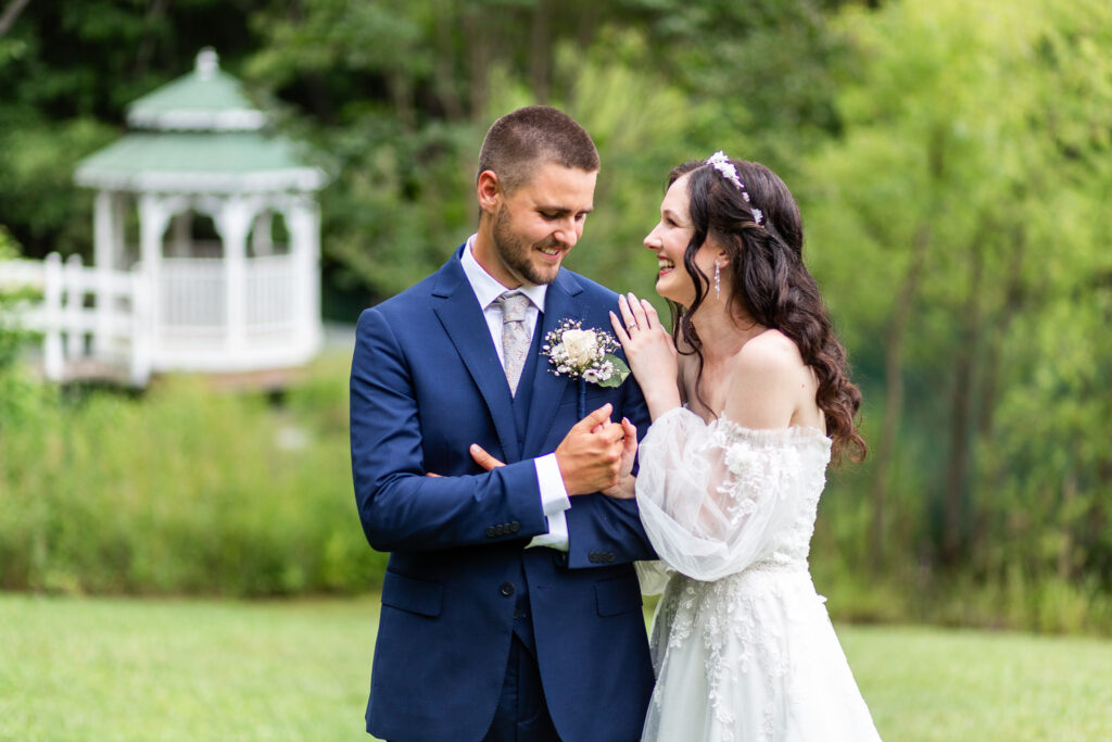 The couple shares a lighthearted moment, smiling at each other in front of the pond and white gazebo, surrounded by lush summer trees—an idyllic Tennessee Mountain Wedding scene.