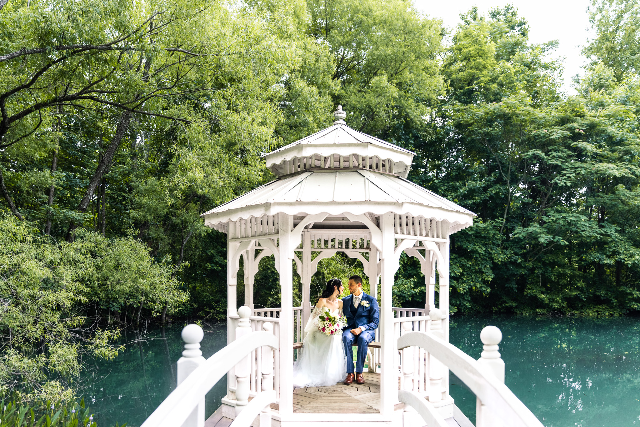 Framed by the soft curves of a white bridge, the couple sits inside the gazebo, gazing at each other and surrounded by serene trees and still waters—an iconic Tennessee Mountain Wedding portrait.