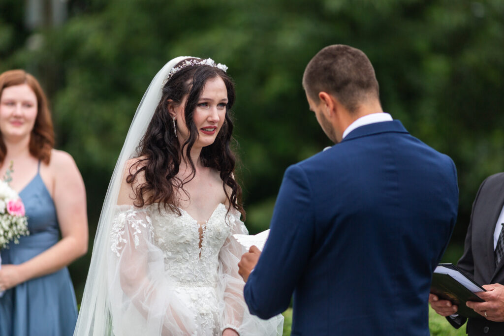 The bride fights back tears while listening to her groom read his vows during their Mountain Willow Manor Wedding ceremony. Her face is filled with emotion as she stands in her lace gown and long veil, holding his words close.