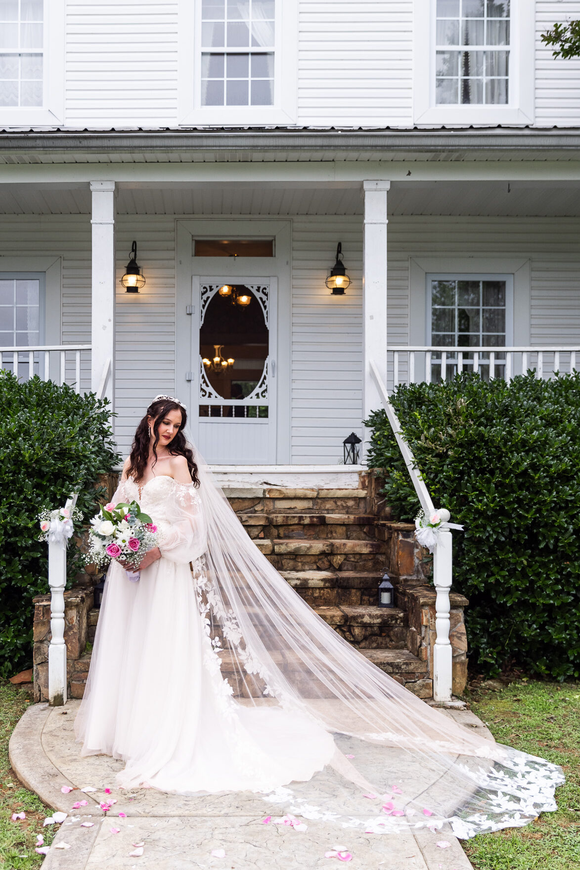 Bride standing alone on the front path of the manor house, her long veil sweeping to the side and floral bouquet in hand. The rustic white paneling and boxwood hedges frame the scene. Chattanooga Wedding Venue.
