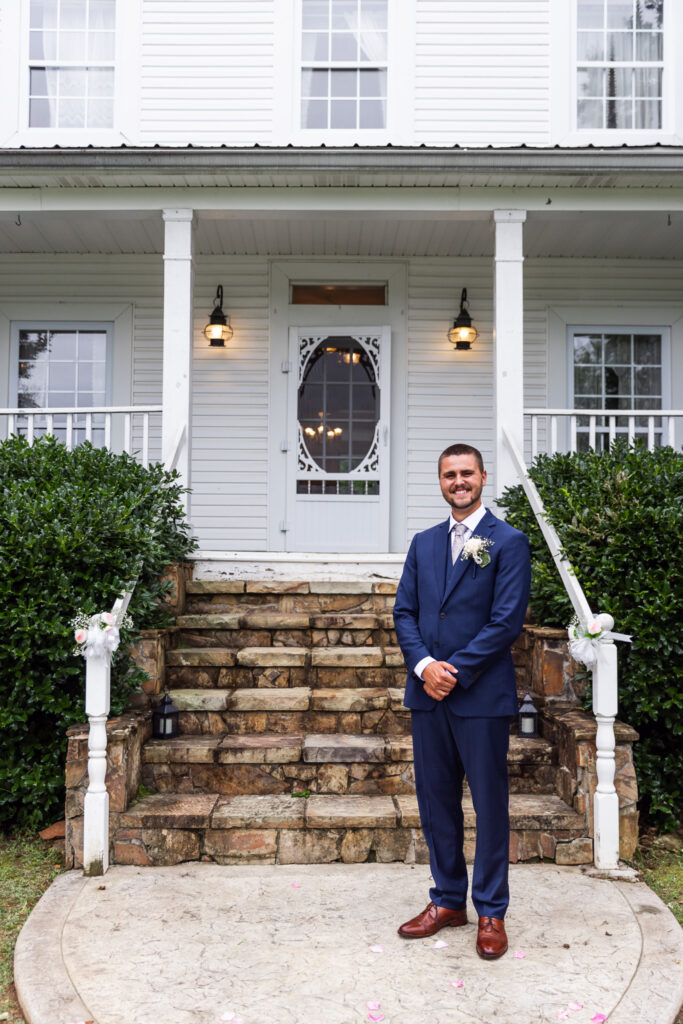 The groom stands smiling on the stone steps in front of the white Victorian-style manor house at Mountain Willow Manor Wedding venue. He’s dressed in a navy suit and boutonniere, ready to welcome guests as soft porch lights glow behind him.