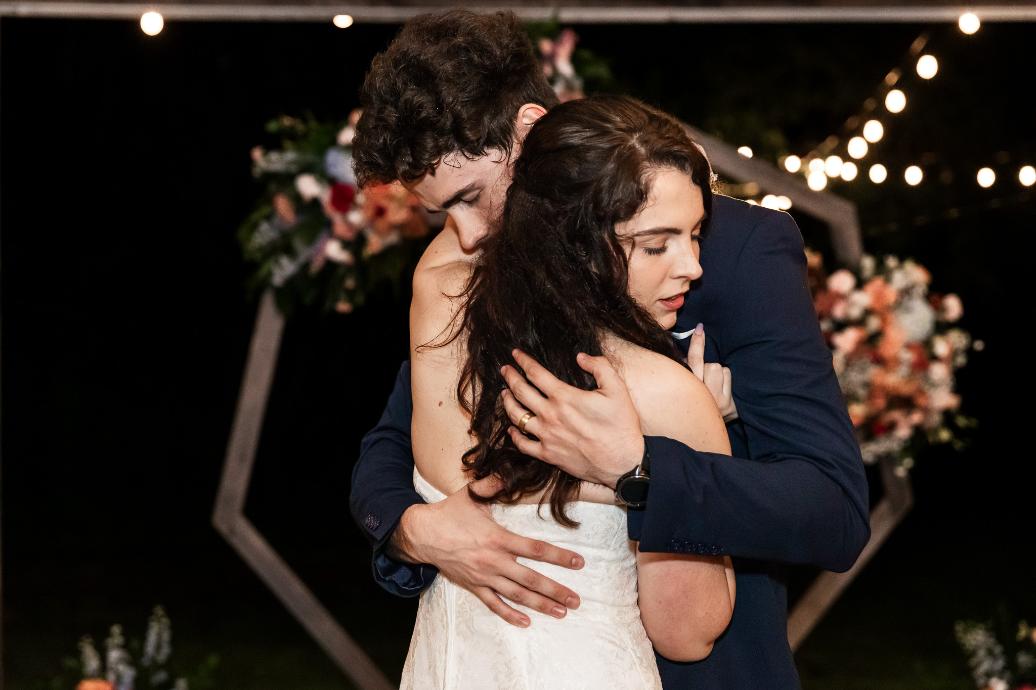 The bride and groom share a heartfelt embrace during their private last dance beneath string lights and a floral hexagon arch at night. The bride closes her eyes, resting her cheek gently on the groom’s shoulder, while his hand wraps tightly around her back. Her lace dress and his navy suit are softly lit by the warm glow of the lights and surrounding florals.