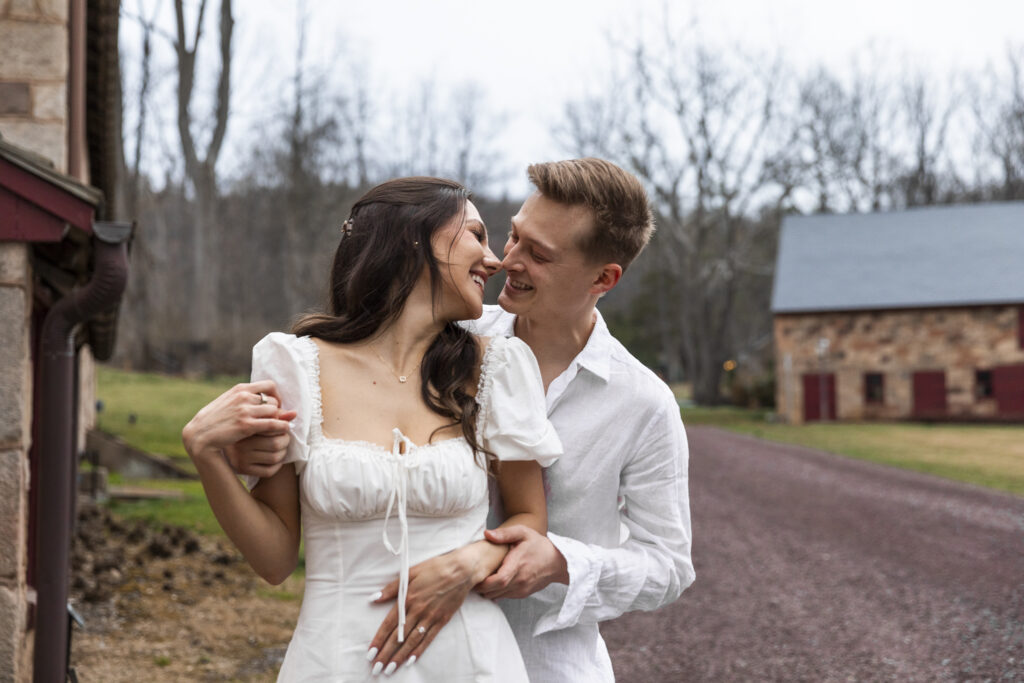 A sweet moment between Alyssa and Noah, nose-to-nose and smiling warmly, captured during their Elizabeth Furnace wedding engagement session with historic stone barns in the distance.