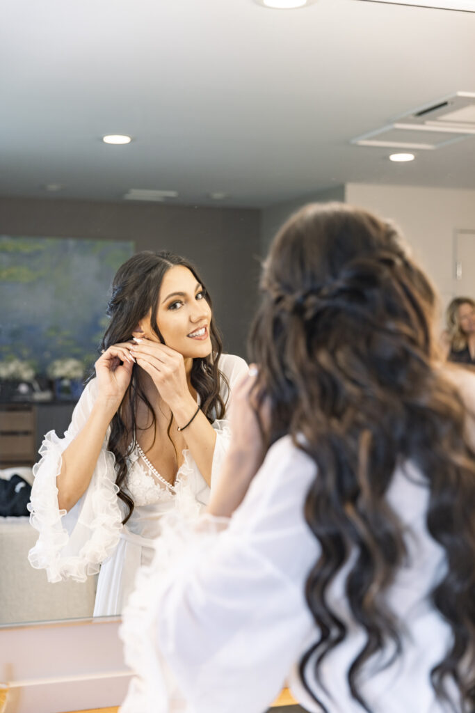 Alyssa smiles in the mirror as she puts on pearl earrings, her reflection glowing with anticipation during the getting ready portion of her Elizabeth Furnace wedding.