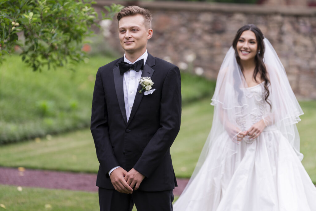 Noah stands in nervous anticipation with a soft smile as Alyssa walks toward him, the stone garden walls of Elizabeth Furnace in the background of this emotional wedding day moment.