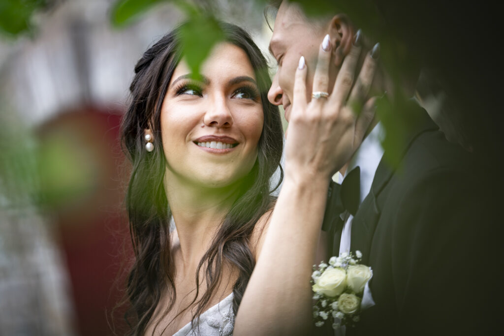 A romantic close-up of the bride cradling her groom’s cheek with her manicured hand, captured through soft green leaves. Her eyes sparkle as she gazes lovingly at him, pearl earrings visible beneath long dark hair. Only a partial view of the groom’s smile and tuxedo is seen as he leans in, emphasizing the bride’s tender expression in this dreamy, hidden garden moment.