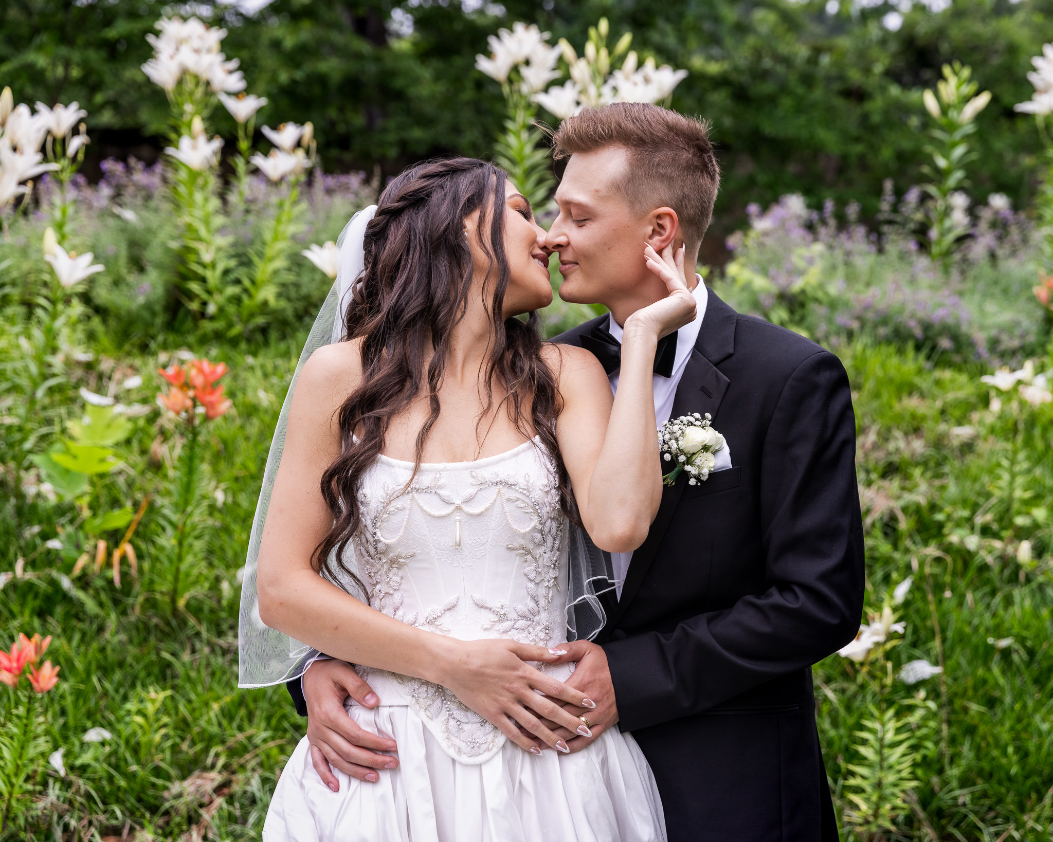 A romantic portrait of Alyssa and Noah kissing gently in the garden, surrounded by lilies and wildflowers—capturing the romance of their Elizabeth Furnace wedding.