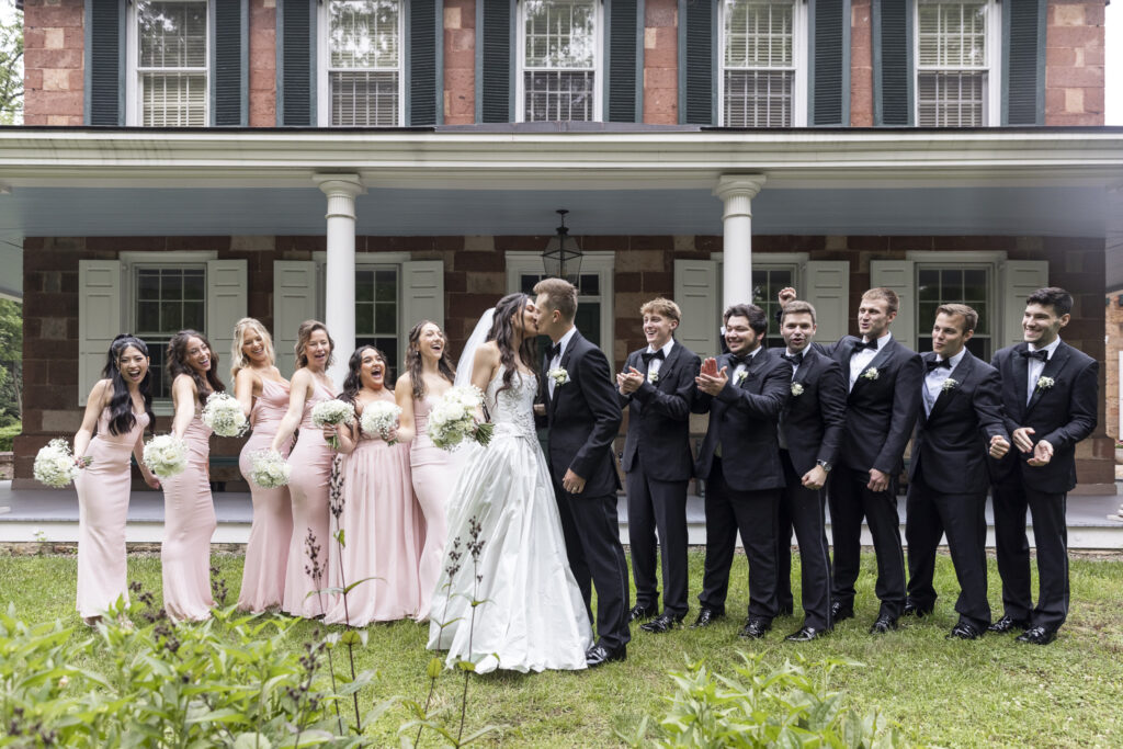 The wedding party— six bridesmaids and six groomsmen—stands grouped in front of a lush garden wall. Everyone is laughing, caught mid-laughter with open smiles and relaxed postures. The bridesmaids’ blush dresses vary slightly in fabric and cut, adding texture, while the groomsmen wear black tuxedos. This Lancaster wedding photo radiates fun and connection.