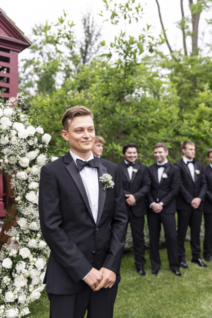 Standing at the altar with his hands gently clasped in front of him, Noah watches his bride approach with a soft, emotional smile. He wears a classic black tuxedo with a crisp white shirt and boutonnière, positioned in front of a floral arch overflowing with white roses and baby’s breath. His groomsmen stand behind him in a staggered row, all in matching attire, as greenery frames the scene. This tender Lancaster wedding photo captures the anticipation and love in the moment before the couple is united.