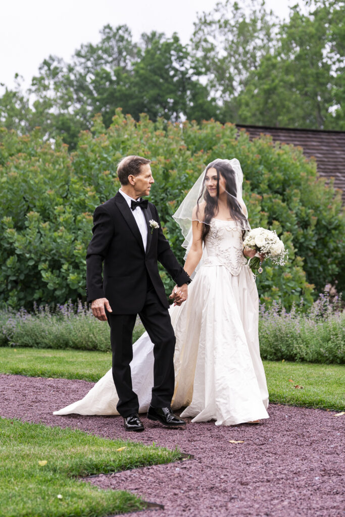 A bride with long dark hair, partially veiled in tulle, walks hand in hand with her father along a red gravel path lined with grass and purple florals. She wears a strapless ivory gown with embroidery at the bodice and carries a bouquet of white flowers. Her father, dressed in a classic black tuxedo, looks at her lovingly. The lush greenery and rustic architecture in the background reflect the charm of a Pennsylvania garden ceremony.