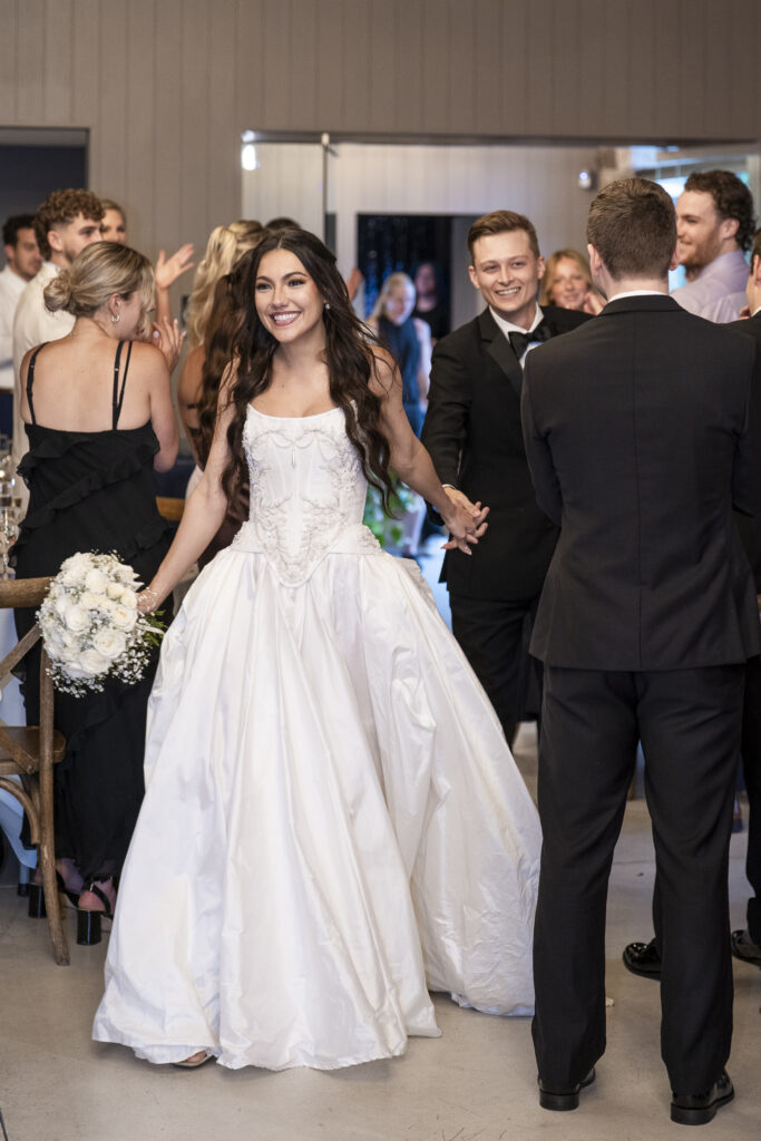 The bride and groom make a grand entrance into their reception, holding hands and smiling as they walk between tables of cheering guests. The bride beams with joy, her white gown shimmering under the indoor lights as she holds a bouquet of white roses. The groom follows just behind, dressed in a black tuxedo with a boutonnière. Guests stand and clap in celebration, creating an atmosphere of excitement and love.