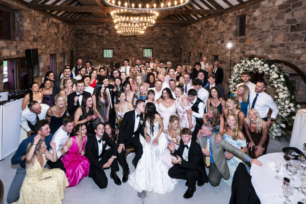 A large group photo taken on the dance floor, featuring the newlyweds seated at the center, sharing a kiss while surrounded by their cheering guests. The room is filled with smiling faces, raised hands, and joyful energy under a series of glowing circular chandeliers and stone walls.