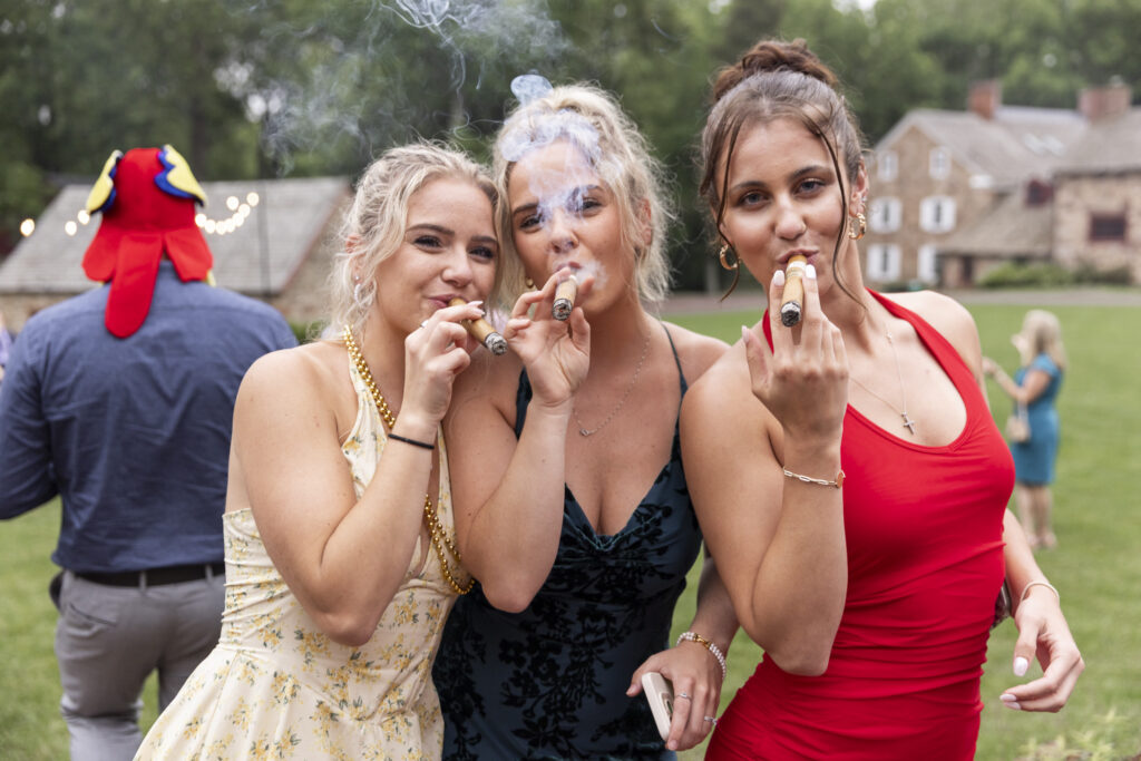 Three women in cocktail dresses—one in yellow floral, one in dark green velvet, and one in red—pose close together outside, each puffing on a cigar and smiling at the camera. Wisps of smoke curl upward, and the relaxed outdoor setting includes a rustic stone house in the background.