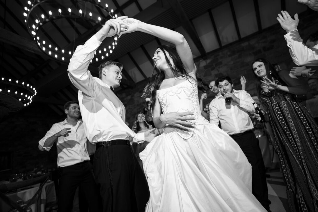 A low, wide-angle black and white photograph of the newlyweds dancing joyfully. The groom, in a white shirt and dark trousers, holds the bride’s hand high as she spins beneath his arm. The bride’s voluminous gown flares out gracefully, her long hair cascading as she smiles up at him. Surrounding guests cheer and clap under large circular chandeliers glowing overhead, adding a celebratory ambiance to the rustic stone-walled reception space.
