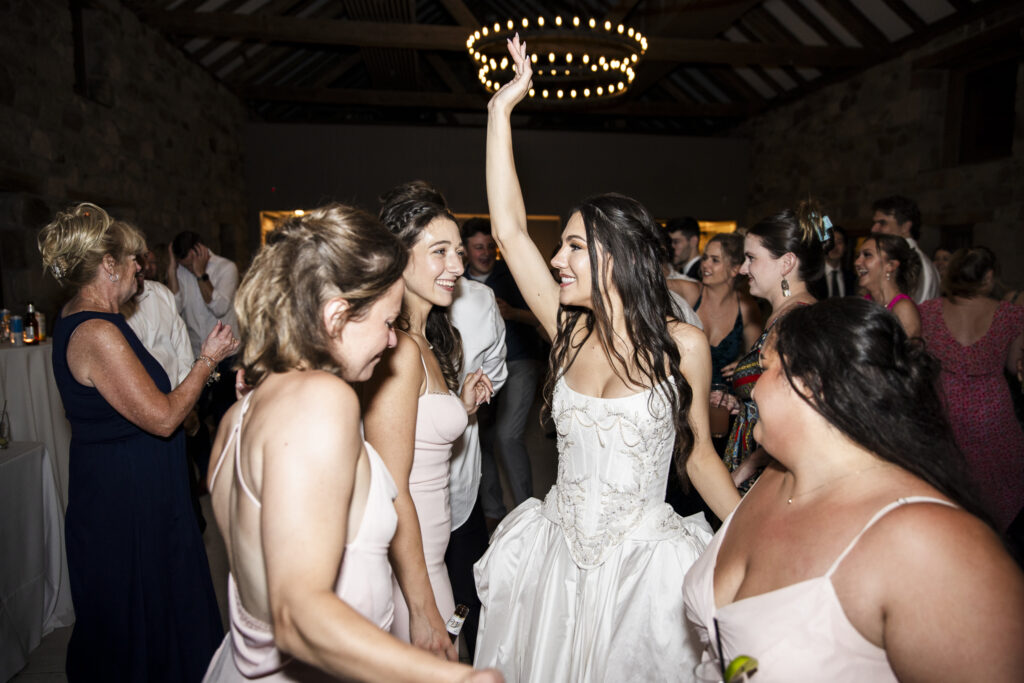The bride, in her intricately beaded gown, raises one arm in the air with a huge smile on her face as she dances surrounded by a lively group of guests. Bridesmaids in blush pink dresses and other attendees in colorful outfits surround her, clapping and cheering beneath warm chandelier light and exposed wooden beams.