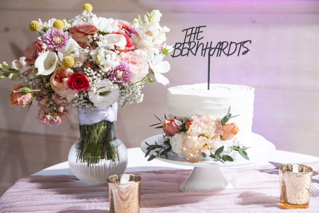 A simple white wedding cake decorated with blush roses and peach flowers sits on a pedestal next to a large vase of matching blooms. The cake topper reads "The Bernhardts," displayed on a table inside a Lancaster wedding venue reception space.