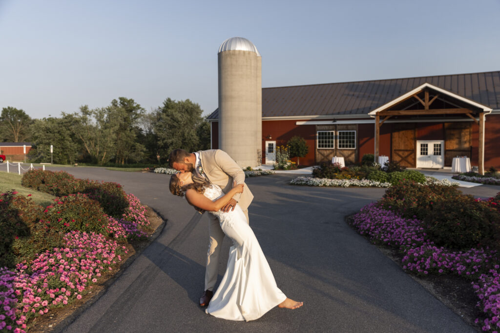 A newlywed couple shares a dramatic kiss at golden hour in front of a red barn with a metal silo, surrounded by vibrant pink and white summer flowers. The groom, wearing a tan suit, leans the barefoot bride into a deep dip as she smiles into the kiss, her white gown flowing to the pavement. The moment is playful, spontaneous, and full of warmth, with soft evening light casting long shadows across the driveway and floral-lined landscaping.