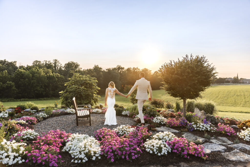 The bride and groom walk hand in hand into the sunset at their Lancaster barn wedding, surrounded by blooming flowerbeds and soft gravel paths. Their backs are to the camera as they stroll through a garden overlook, bathed in golden light.