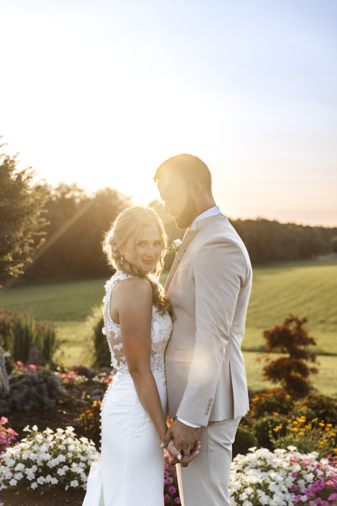 The bride and groom stand in a garden bathed in golden light, holding hands and facing forward with soft smiles. The bride turns slightly toward the camera, her long curls and lace gown glowing as the sun sets behind them, casting warm light across the field and surrounding trees.