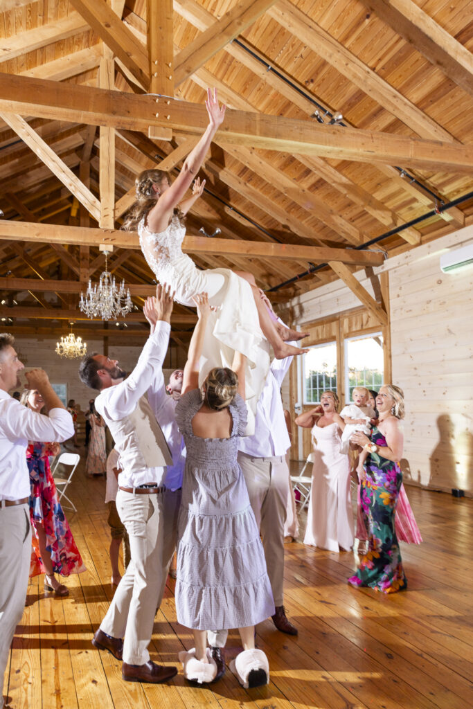 The bride is lifted high into the air by guests during a joyful moment at her Lancaster barn wedding reception. Her arms are outstretched as she laughs, surrounded by friends beneath exposed beams and glowing chandeliers in the rustic wood venue.