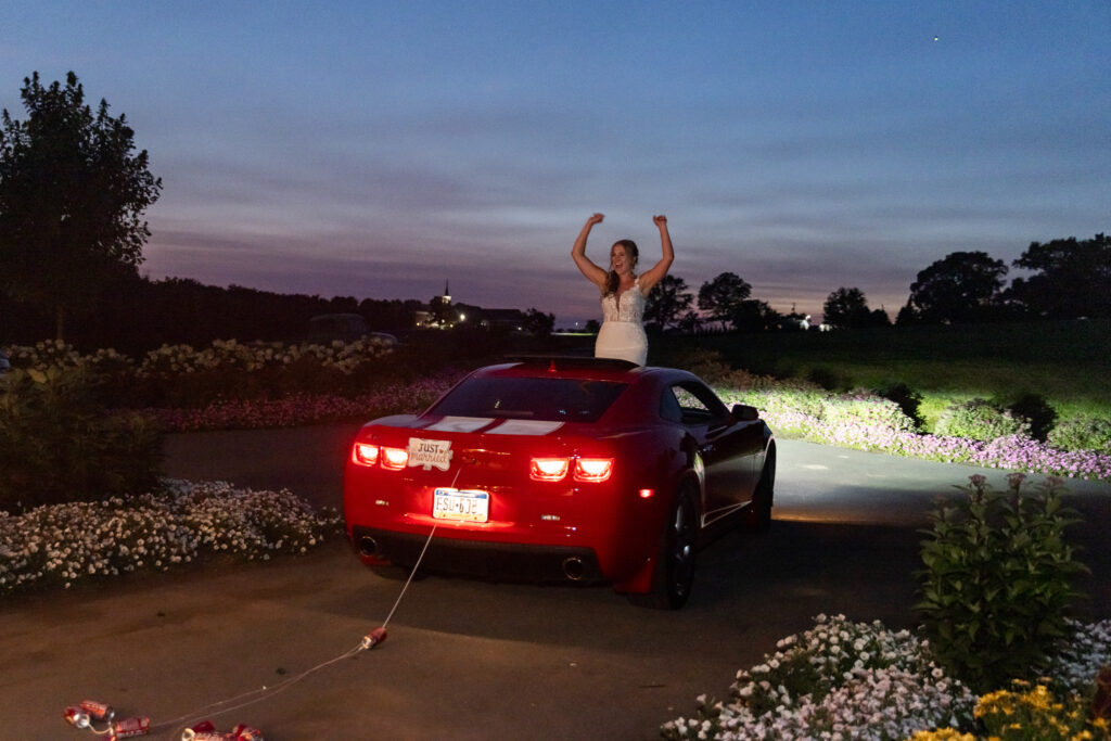 The bride stands tall through the sunroof of a red sports car adorned with a "Just Married" sign, waving her arms in celebration as she and her groom drive off from their Lancaster barn wedding. Tin cans trail behind the car as dusk falls over rows of flowers and trees.