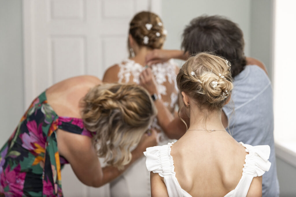 From behind, a flower girl watches as two women help button the bride into her dress. The bride’s hair is styled in an elegant updo adorned with pearls and flowers, mirrored in the young girl’s similar hairstyle. The group is gathered tightly in a calm getting-ready room.
