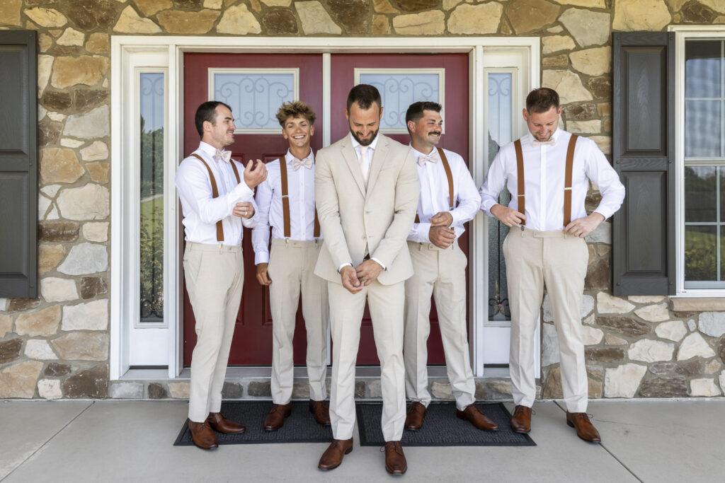The groom stands in front of a red door with four groomsmen dressed in coordinating tan pants, white shirts, and brown suspenders. They laugh and adjust their outfits as the groom grins, all gathered on a shaded porch with stone walls, exuding laid-back energy and friendship.
