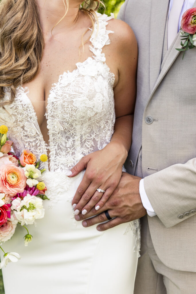 A close-up shot of the couple’s hands resting on the bride’s waist, showcasing both wedding bands and the intricate lace of her gown. The bouquet peeks into the frame, adding a pop of color that ties into the floral decor of this Lancaster wedding venue.