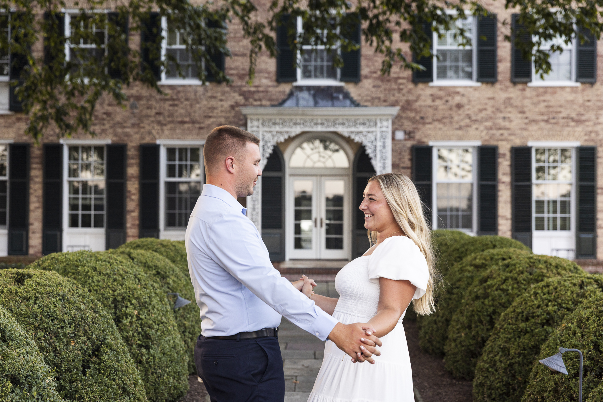 Close-up of the couple smiling and looking into each other’s eyes in front of the grand doorway at Drumore Estate. Man wears a light blue shirt and dark slacks; woman wears a flowing white dress with puff sleeves, her blonde hair cascading down her back.
