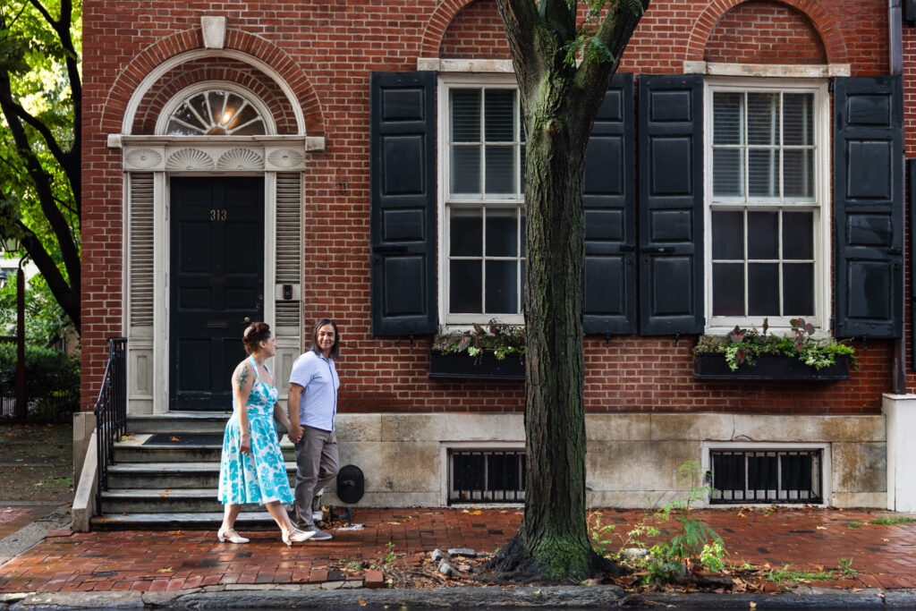 A couple walks hand in hand down a brick sidewalk in front of a historic red brick townhouse with black shutters and white trim. The woman in a turquoise floral dress looks at her partner, who smiles warmly back at her. A large tree divides the frame, grounding the scene in the charm of old Philadelphia architecture — a classic example of Philadelphia Engagement Photos.