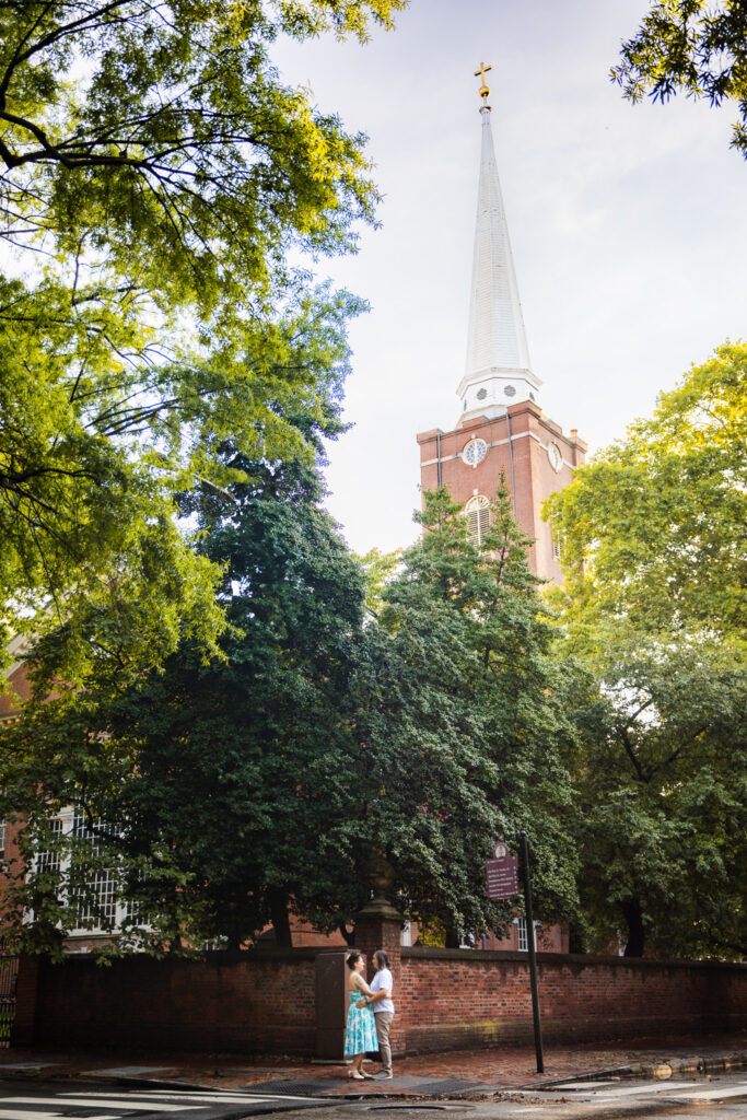 A couple stands on a cobblestone sidewalk beneath a canopy of lush green trees, framed by the towering white steeple of Saint Peters Church in the background. The woman wears a turquoise patterned dress, and the man is in a light shirt and khaki pants. They hold each other close at the edge of the scene, adding a tender, human scale to the grand architecture towering above them.