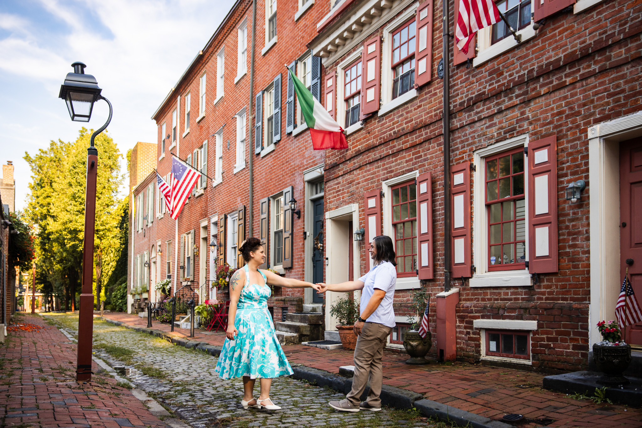 On a cobblestone street lined with colonial row houses, the couple reaches across the space between them to clasp hands. Flags wave from the windows, and the afternoon sun casts a golden glow, capturing the sense of place that makes Philadelphia engagement photos so timeless.