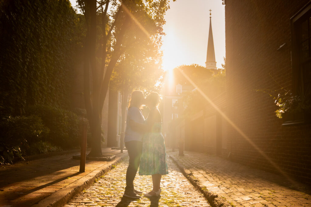 The couple shares a kiss in a narrow brick alleyway as the golden sun sets directly behind them. Their silhouettes glow with rim light as sunbeams streak through the trees and reflect on the cobblestones. In the distance, the Saint Peter's Church steeple rises into the sky, completing the timeless, romantic backdrop of these Philadelphia engagement photos