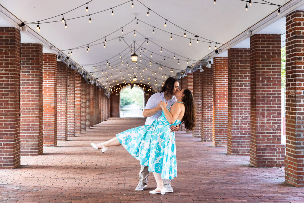 Beneath a vaulted brick arcade glowing with rows of string lights, he dips her back into a kiss as her turquoise dress flares around her. The repeating arches and soft lights frame them in a way that feels cinematic, a moment that could only belong to Philadelphia engagement photos.