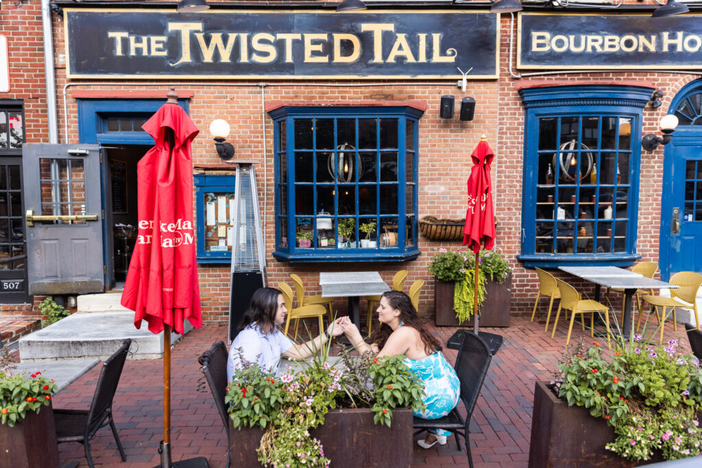 Seated at an outdoor café table surrounded by planters overflowing with flowers, the couple holds hands and laughs together. The brick backdrop and bold blue trim of the restaurant windows highlight the city’s character, adding a vibrant layer to their Philadelphia engagement photos outside of The Twisted Tail