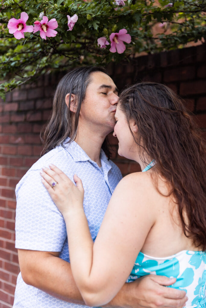 The man presses a kiss to the woman’s forehead as she smiles with her eyes closed, leaning into him under a branch of blooming pink flowers. They stand in front of a red brick wall, her hand resting against his chest, the engagement ring clearly visible.