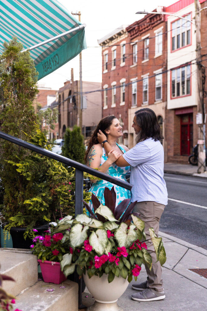 The man gently tucks the woman’s hair behind her ear as they share a laugh beside a storefront with striped green-and-white awnings. Planters overflowing with bright pink flowers frame the foreground, adding color and charm to this South Street moment.
