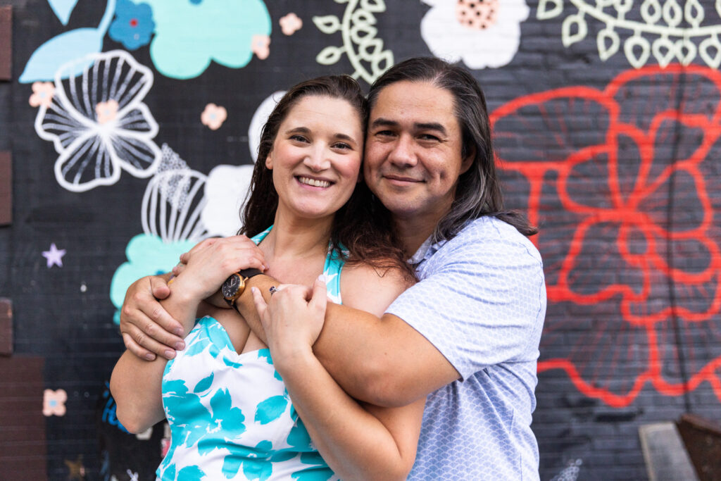 Wrapped in each other’s arms, the couple stands in front of a bold floral mural painted in shades of teal, red, and white. Their joyful smiles and affectionate embrace capture a warm, genuine moment in the heart of South Street.