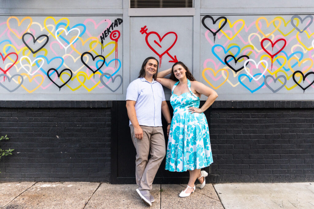 The couple poses in front of a colorful mural covered with overlapping heart outlines in bright shades of yellow, red, blue, and black. They lean casually against the wall, smiling with relaxed confidence, embodying the playful spirit of South Street’s vibrant art scene.