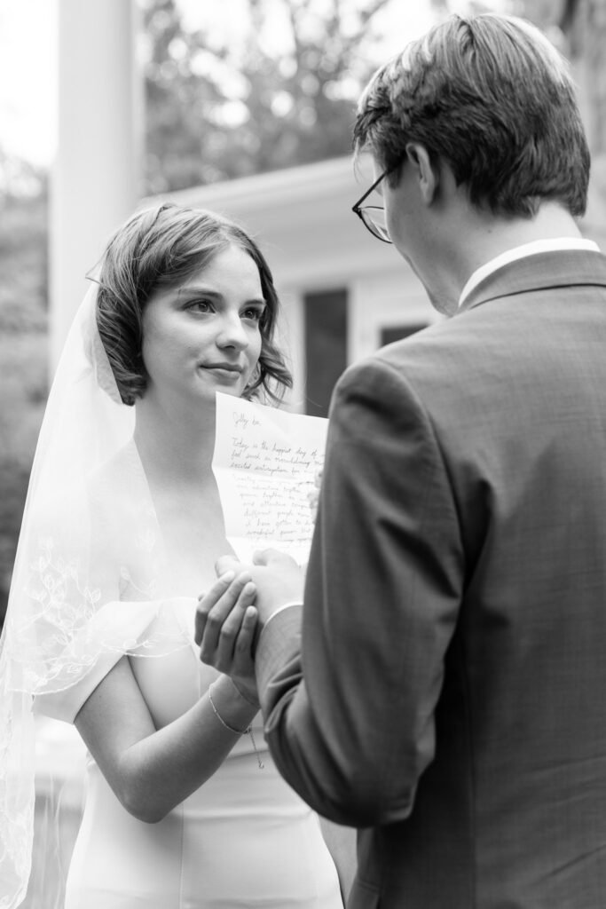 Black and white image of the groom reading handwritten vows to the bride during their first look at McCoy Farms, she smiles as tears well in her eyes and her veil drapess softly over her shoulders.
