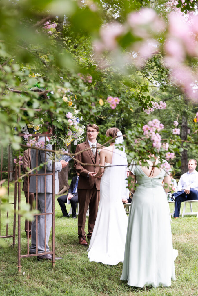 Romantic view of the McCoy Farms Wedding Ceremony framed through pink blossoms, capturing the couple standing with the officiant and wedding party.