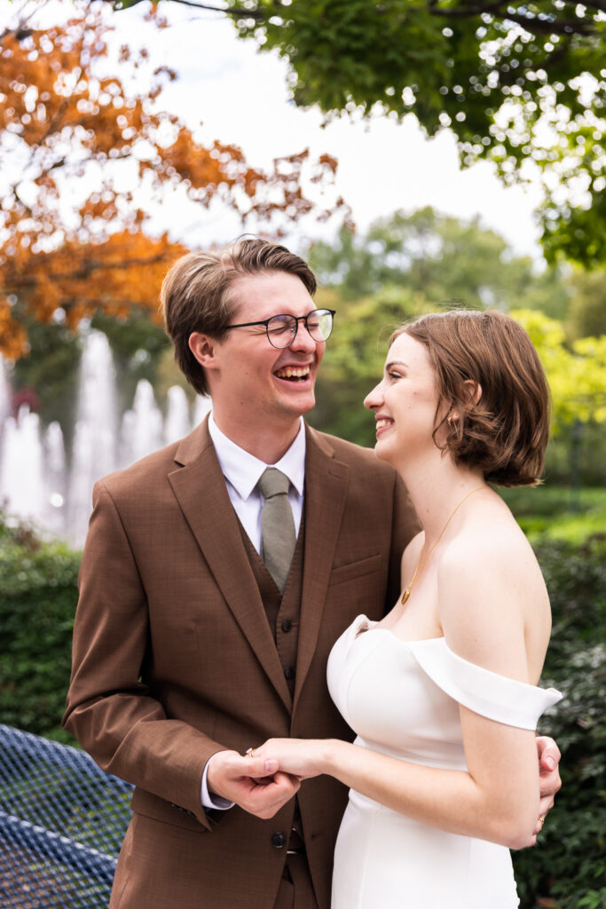 The bride and groom laugh together in a garden, holding hands, with fountains spraying in the blurred background and autumn-colored leaves contrasting against the greenery.