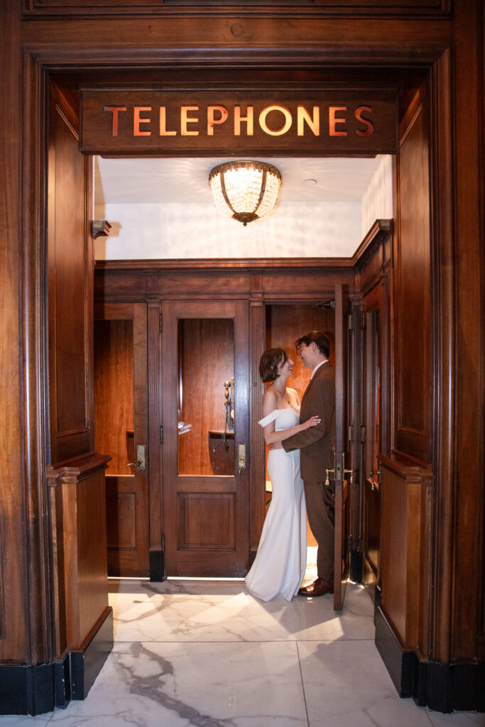 Bride and groom smiling at each other while standing in the classic wood phone booth area of their Read House Wedding venue, beneath a sign reading “Telephones.”