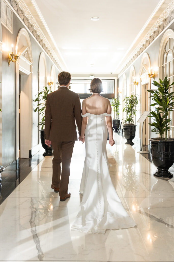 A bride and groom walk hand in hand down a bright, elegant hallway as sunlight spills in, as they head to their Read House wedding reception. The deco building features marble floors, tall potted plants, and ornate wall sconces.
