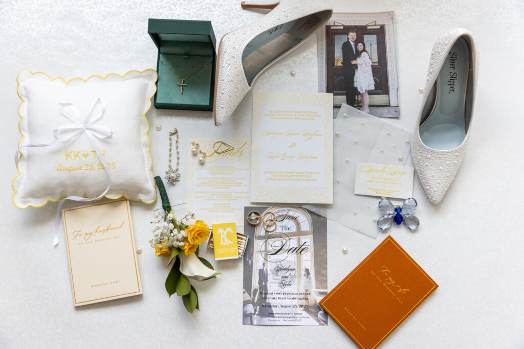 A flat lay arrangement of wedding day details displayed on a soft patterned background, featuring white beaded heels, a gold and ivory wedding invitation suite, pearl-accented jewelry, a green box holding a cross necklace, vow books for the bride and groom, and a ring bearer pillow embroidered with the couple’s initials and wedding date. A boutonniere with yellow roses and calla lilies rests nearby alongside a framed engagement photo of the couple.