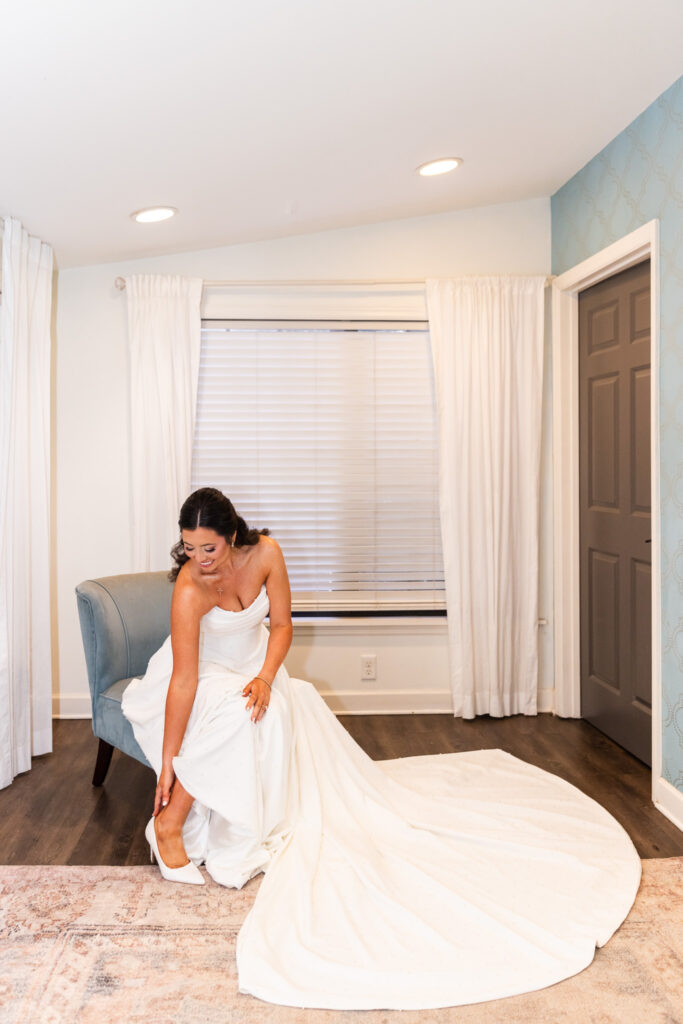 The bride, wearing a strapless white gown with a long flowing train, sits gracefully in a soft blue chair as she slips on her white heels. She looks down with a joyful smile, her veil cascading over the chair.