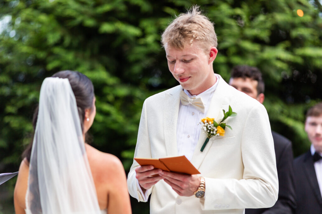 Standing in a white suit and gold bowtie, the groom gazes down at a small orange vow book, reading with tenderness as the bride listens quietly with her veil cascading down her back. Behind them, groomsmen in dark suits and tall trees frame the setting at one of the outdoor wedding venues in Chattanooga, where the vows felt deeply personal and heartfelt