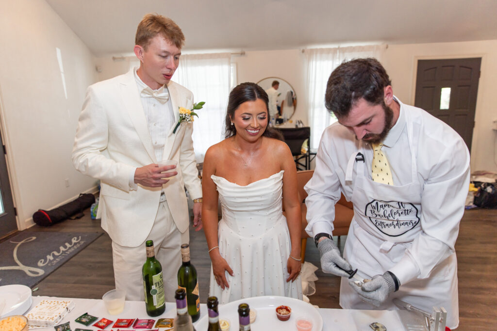 The bride and groom stand side by side indoors, grinning as the bride’s brother leans over a tray to shuck fresh oysters just for them. The bride’s pearl-dotted dress glimmers under soft light, and bottles of wine line the table in front of them, creating an intimate, playful moment shared privately with the bridal party.