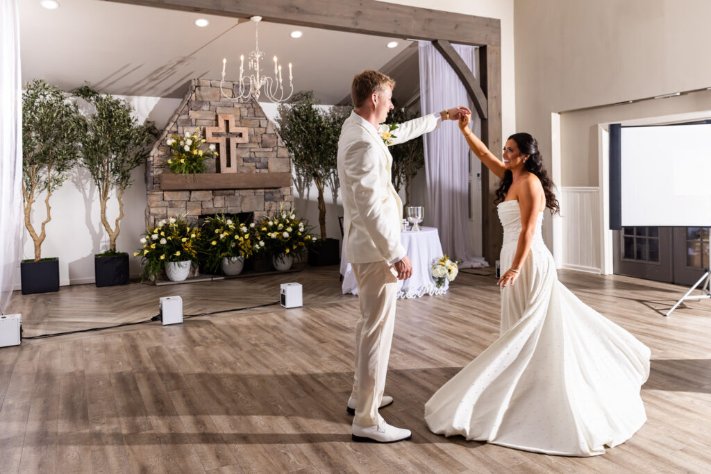 On the wooden dance floor of their reception hall, the groom lifts the bride’s hand to twirl her, sending her pearl-detailed gown fanning outward in a graceful circle. Behind them, a stone fireplace decorated with yellow and white flowers and tall greenery provides a warm backdrop under a sparkling chandelier.