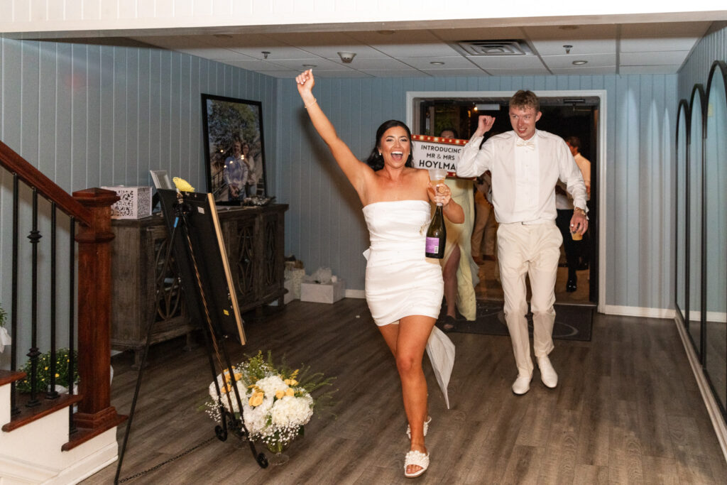 The bride, now in her reception dress, raises her arm in celebration while holding a champagne bottle, followed by the groom as they make their lively entrance into The Venue Chattanooga’s reception space.